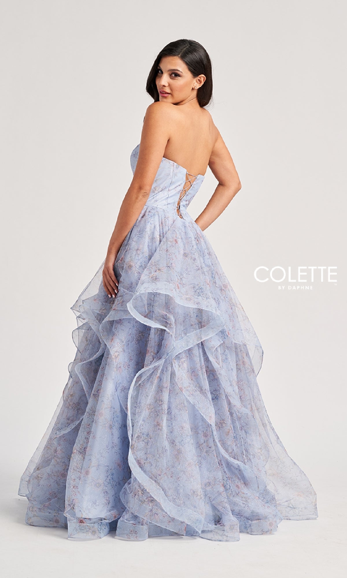 A woman poses with her back mostly turned, looking over her shoulder in the Colette CL8180 long prom dress, featuring a strapless design and layered light purple floral pattern.